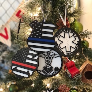 May include: Four Christmas ornaments shaped like badges for police, firefighters, EMTs, and a medical symbol. The ornaments are black, white, red, and blue with the American flag on each.
