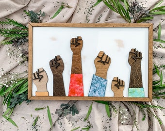 May include: A wooden wall art piece featuring five raised fists in different colors, including brown, red, blue, and green. The fists are arranged in a row, with the red fist in the center.
