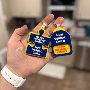 May include: Two keychains with the text "NON VERBAL CHILD" and "TAP FOR EMERGENCY CONTACT". One keychain is a blue puzzle piece shape with yellow accents. The other is a blue tag with yellow accents. Both have a silver key ring.