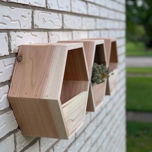 May include: Three wooden hexagonal shelves mounted on a white brick wall. The shelves are unfinished and have a natural wood color. The shelves are arranged in a row, with the largest shelf on the left and the smallest shelf on the right.