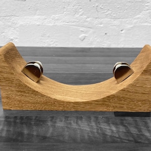 May include: A wooden stand with a curved shape, designed to hold two small, silver-coloured objects. The stand is made of light brown wood and has a smooth finish.
