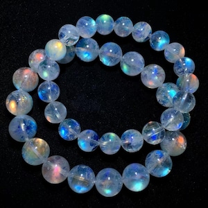 May include: A circular bracelet composed of translucent, spherical moonstone beads. Each bead displays a distinct adularescence, reflecting internal flashes of blue, orange, and white light against a dark background, arranged in a continuous loop.