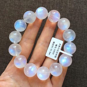 May include: A bracelet composed of round, translucent moonstone beads with a blue iridescent sheen. A white rectangular tag is attached to the bracelet, featuring the text: 天然月光石手链, 编号: F0932, 珠径约: 15.5mm.