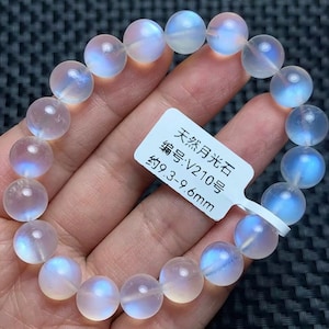 May include: A bracelet made of round, translucent moonstone beads with a blue iridescent sheen. A white rectangular tag is attached to the bracelet, featuring the text: 天然月光石, 编号:V210号, 约9.3-9.6mm. The beads are strung on a clear elastic cord.
