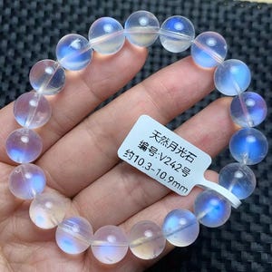 May include: A bracelet composed of translucent, spherical moonstone beads exhibiting a blue adularescence. A white rectangular tag is attached, featuring the text "天然月光石 编号:V242号 约10.3-10.9mm", indicating a bead diameter of approximately 10.3 to 10.9 mm.