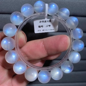 May include: A circular bracelet composed of smooth, translucent, spherical beads with a blue iridescent sheen. A white rectangular tag is attached, featuring the text "冰体蓝月光", "编号: J2号", and "尺寸 约11.5 11.8mm", indicating a bead size of approximately 11.5 to 11.8 mm.
