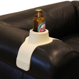 May include: A white plastic beverage holder that attaches to the arm of a couch. The holder has a raised lip to keep a bottle or can from tipping over. A brown glass bottle with a green label is in the holder.
