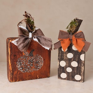 May include: Two rectangular wooden blocks shaped as pumpkins. The left block is stained orange with a leopard print pumpkin graphic and a brown and white fabric bow. The right block is stained dark brown with white polka dots and an orange bow.