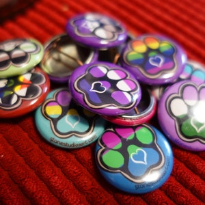 May include: A collection of colorful, round buttons with various paw print designs and heart accents. The buttons feature a range of colors, including purple, blue, green, and red, with some displaying rainbow patterns. The text "stonestudioart.com" is visible on some buttons.