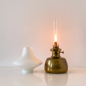 May include: A brass oil lamp with a white glass shade and a lit wick. The lamp is on a white surface.