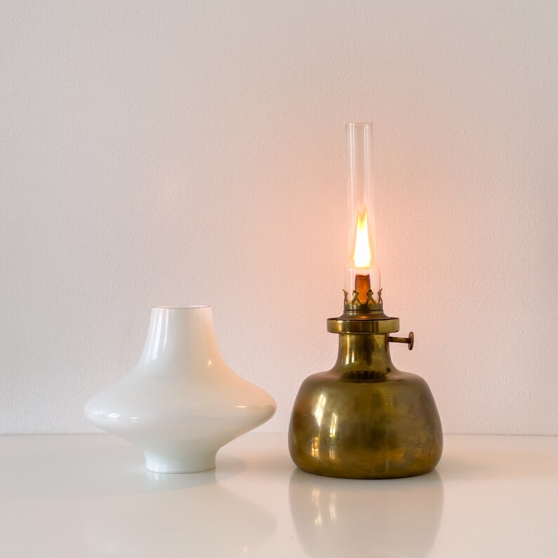 May include: A brass oil lamp with a white glass shade and a lit wick. The lamp is on a white surface.