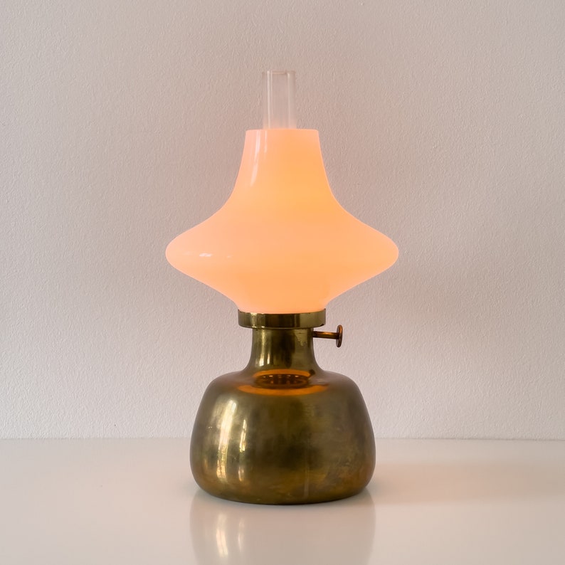 May include: A brass table lamp with a white glass shade. The shade is shaped like a wide, flat bowl with a small opening at the top. The lamp is lit and casting a warm glow.