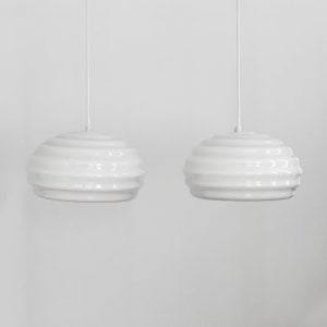 May include: Two white pendant lights with a wavy, ribbed design hang from white cords against a white background.