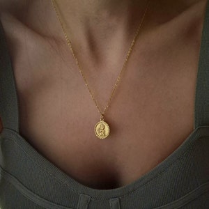 Op de afbeelding: Een gouden ketting met een ronde hanger met een gedetailleerd beeld van een cherubijn. De hanger heeft een touwachtige rand.
