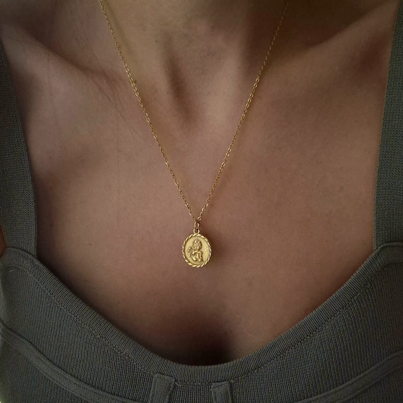 Op de afbeelding: Een gouden ketting met een ronde hanger met een gedetailleerd beeld van een cherubijn. De hanger heeft een touwachtige rand.