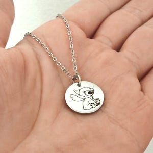 May include: A silver-colored necklace featuring a round pendant with an engraved image of a cartoon character. The pendant is attached to a delicate chain. The necklace is held in a person's hand.