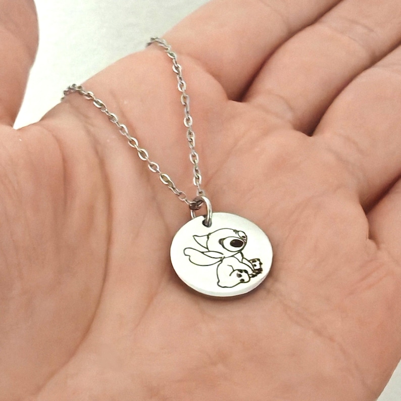 May include: A silver-colored necklace featuring a round pendant with an engraved image of a cartoon character. The pendant is attached to a delicate chain. The necklace is held in a person's hand.