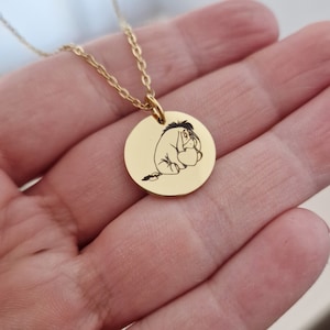 Op de afbeelding: Een goudkleurige ketting met een ronde hanger. De hanger heeft een zwarte illustratie van Iejoor uit Winnie de Poeh. De hanger is bevestigd aan een fijne gouden ketting. Een eenvoudig en elegant ontwerp. Een charmant accessoire.