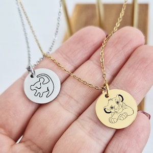 Könnte beinhalten: Zwei goldene Halsketten mit runden Anhängern. Ein Anhänger zeigt eine schwarze Umrandung von Simba aus dem König der Löwen. Der andere Anhänger zeigt eine Schwarzweißillustration von Simba als Jungtier.