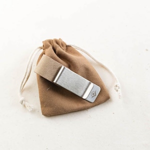 Puede incluir: Un clip de dinero plateado con un pequeño logotipo en la parte delantera, descansando dentro de una bolsa de tela marrón con cordón.