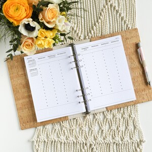 Könnte beinhalten: Ein weißer Planer mit einem Korkbezug und einem Blumenarrangement darauf. Der Planer hat ein Ringbindungssystem und ist auf eine Monatskalenderansicht geöffnet. Der Kalender ist in Spanisch und umfasst die Monate Januar und Februar.