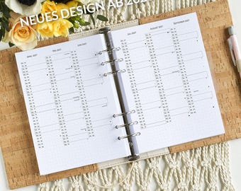 May include: A white planner with a cork cover and a silver binder. The planner has a calendar layout for the months of April through September 2027. The text "NEUES DESIGN AB 2026!" is printed on the cover.