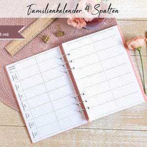 Könnte beinhalten: Ein rosa A5 (14,8 x 21 cm) Familienkalender mit vier Spalten. Der Kalender ist auf einer leeren Seite mit einem gepunkteten Raster geöffnet. Der Kalender liegt auf einem Holztisch mit einer rosa Unterlage und einem Lineal.