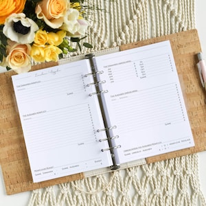 Könnte beinhalten: Offener Finanzplaner mit weißen Seiten, mit Abschnitten für Einnahmen und Ausgaben. Der Planer hat einen Korkbrett-Einband. Ein Stift liegt daneben, und Blumen sind im Hintergrund. Der Planer ist geöffnet.