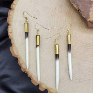 May include: A set of three long, white and black earrings with gold accents. The earrings are made of bone and have a gold-colored metal top.