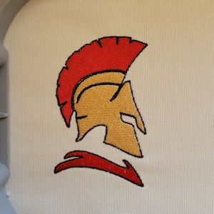 Puede incluir: Diseño bordado de un casco espartano dorado con una pluma roja. El casco está mirando hacia la derecha y tiene un diseño estilizado.