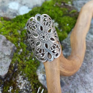May include: A silver filigree ring with a detailed floral design. The ring is on a wooden stick.