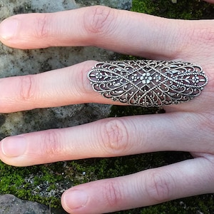 May include: A silver filigree ring with a delicate floral design. The ring is wide and sits on the middle finger.
