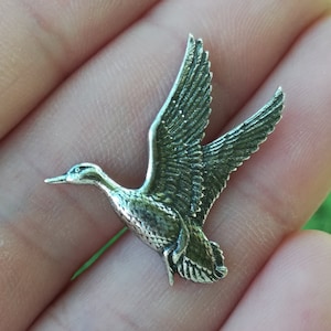 Peut inclure: Breloque en métal argenté en forme de canard avec des ailes détaillées et un design réaliste. Le canard est en vol, les ailes déployées.