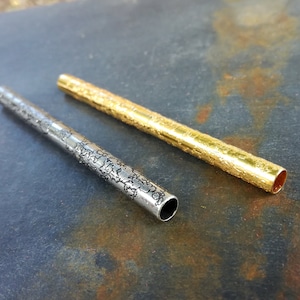 May include: Two metal tubes, one gold and one silver, with a textured surface. The tubes are lying on a dark gray surface.