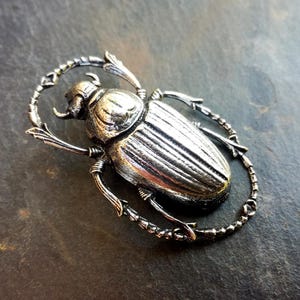 Könnte beinhalten: Silberfarbene Metall-Skarabäus-Brosche. Der Käfer ist mit strukturierten Flügeln und Beinen versehen, die in einem ovalen Rahmen angeordnet sind. Die Brosche hat ein antikes Finish, das ihr einen Vintage-Look verleiht. Das Design ist von altägyptischem Schmuck inspiriert.