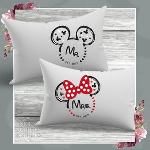 May include: Two white decorative cushions with Mickey Mouse-themed designs. One cushion has a black outline with "Mr." and "Est. 2025." The other features a red polka-dot bow and "Mrs." with the same text. The cushions are on a wooden surface.