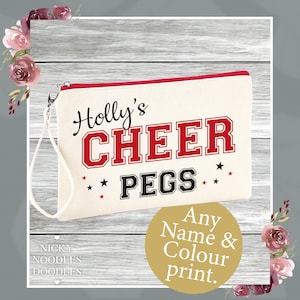 Può includere: Pochette con cerniera color crema con la scritta "Holly's CHEER PEGS" in rosso e nero, con piccole stelle nere. La pochette ha una cerniera rossa e un cinturino da polso. Un cerchio dorato dice "Any Name & Colour print."