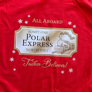 May include: A red t-shirt featuring a gold and white design with the text "All Aboard" and "Admit One Polar Express North Pole." The design includes a train illustration and the phrase "Tucker Believes!" with snowflake accents.