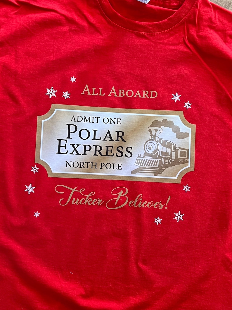 May include: A red t-shirt featuring a gold and white design with the text "All Aboard" and "Admit One Polar Express North Pole." The design includes a train illustration and the phrase "Tucker Believes!" with snowflake accents.