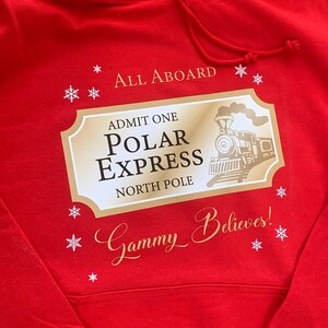 May include: A red hooded sweatshirt with a gold and white graphic. The graphic reads "All Aboard" and features a Polar Express ticket design with a train illustration and the words "Polar Express North Pole". The text "Gammy Believes!" is also included.