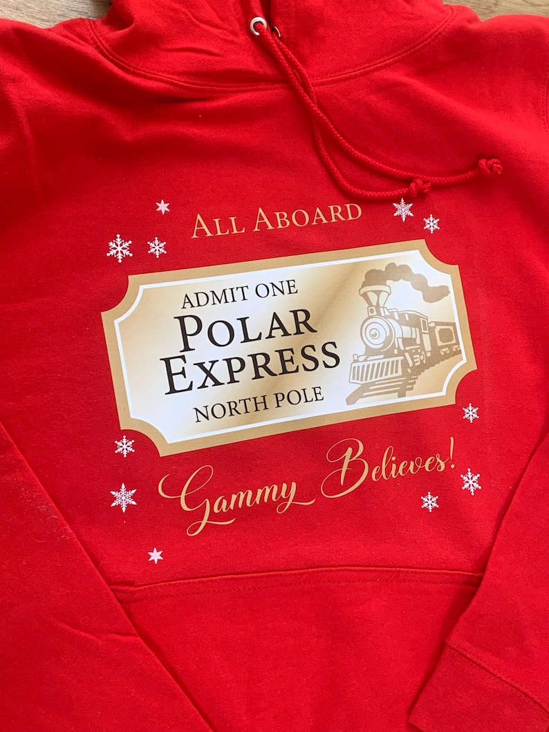 May include: A red hooded sweatshirt with a gold and white graphic. The graphic reads "All Aboard" and features a Polar Express ticket design with a train illustration and the words "Polar Express North Pole". The text "Gammy Believes!" is also included.