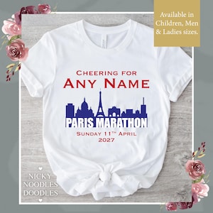 Puede incluir: Camiseta blanca con un diseño para el Maratón de París 2027. El diseño incluye el texto "CHEERING FOR ANY NAME" sobre una silueta azul de los monumentos de París, y la fecha "DOMINGO 11 DE ABRIL DE 2027".
