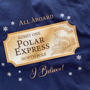May include: Navy blue t-shirt featuring a gold ticket design with a train illustration and the words "POLAR EXPRESS" and "NORTH POLE". The shirt also has the phrases "ALL ABOARD" and "I Believe!" with snowflake accents.