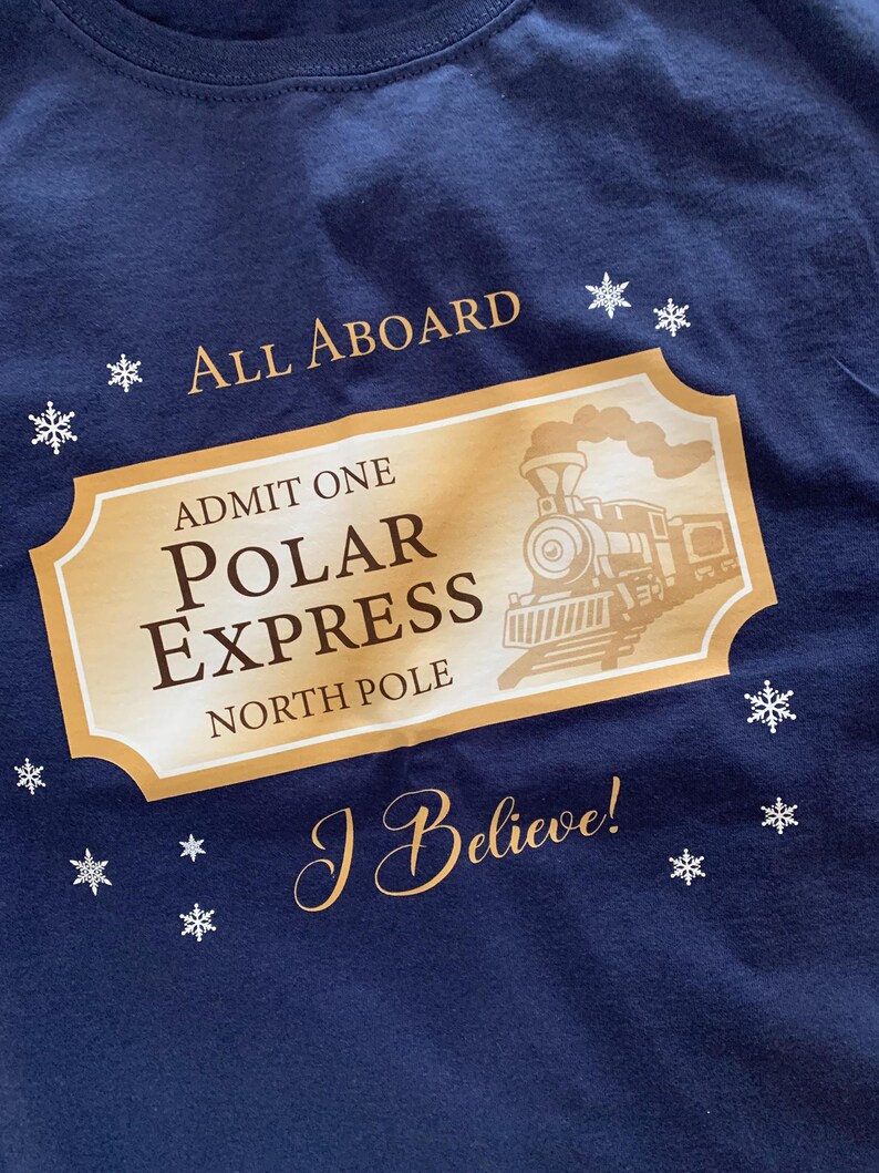 May include: Navy blue t-shirt featuring a gold ticket design with a train illustration and the words "POLAR EXPRESS" and "NORTH POLE". The shirt also has the phrases "ALL ABOARD" and "I Believe!" with snowflake accents.