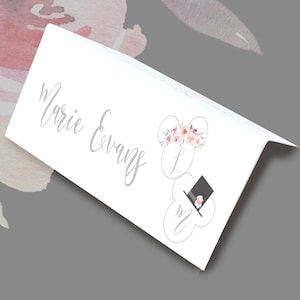 May include: A white folded place card with the name "Marie Evans" in gray script. The card features floral and hat illustrations. The floral illustration is pink and white. The hat illustration is black with a pink flower.