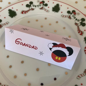 Könnte beinhalten: Weiße Tischkarte mit einer roten und schwarzen Mickey Maus, die einen Weihnachtsmann-Hut trägt. Die Tischkarte trägt den Schriftzug "GRANDAD" in roter, kursiv geschriebener Schrift. Die Tischkarte ist mit Schneeflocken verziert.