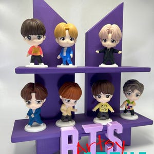 May include: A purple display shelf featuring seven small figurines of cartoon characters. The shelf is shaped like a stylized letter M, with the letters "BTS" and "Artsy Geeks Family" in front. The figurines are wearing various outfits.