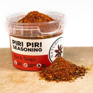 May include: A clear plastic container filled with a red and brown spice blend labelled "Piri Piri Seasoning Hot & Tangy". The label also includes the text "Nature" and a flower logo. The container is sitting on a brown surface with a small pile of the spice blend in front of it.