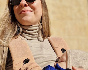 Puede incluir: Una mujer con gafas de sol negras y un cuello alto beige, sosteniendo a un bebé en un portabebés. El bebé lleva un gorro gris y está envuelto en tela azul y marrón. El portabebés es beige con cierres negros.