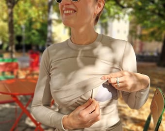 Puede incluir: Una camiseta beige de manga larga con cuello redondo y una discreta abertura para la lactancia. La persona de la imagen lleva vaqueros azules y gafas de sol redondas. El fondo es un parque con árboles y mesas rojas.